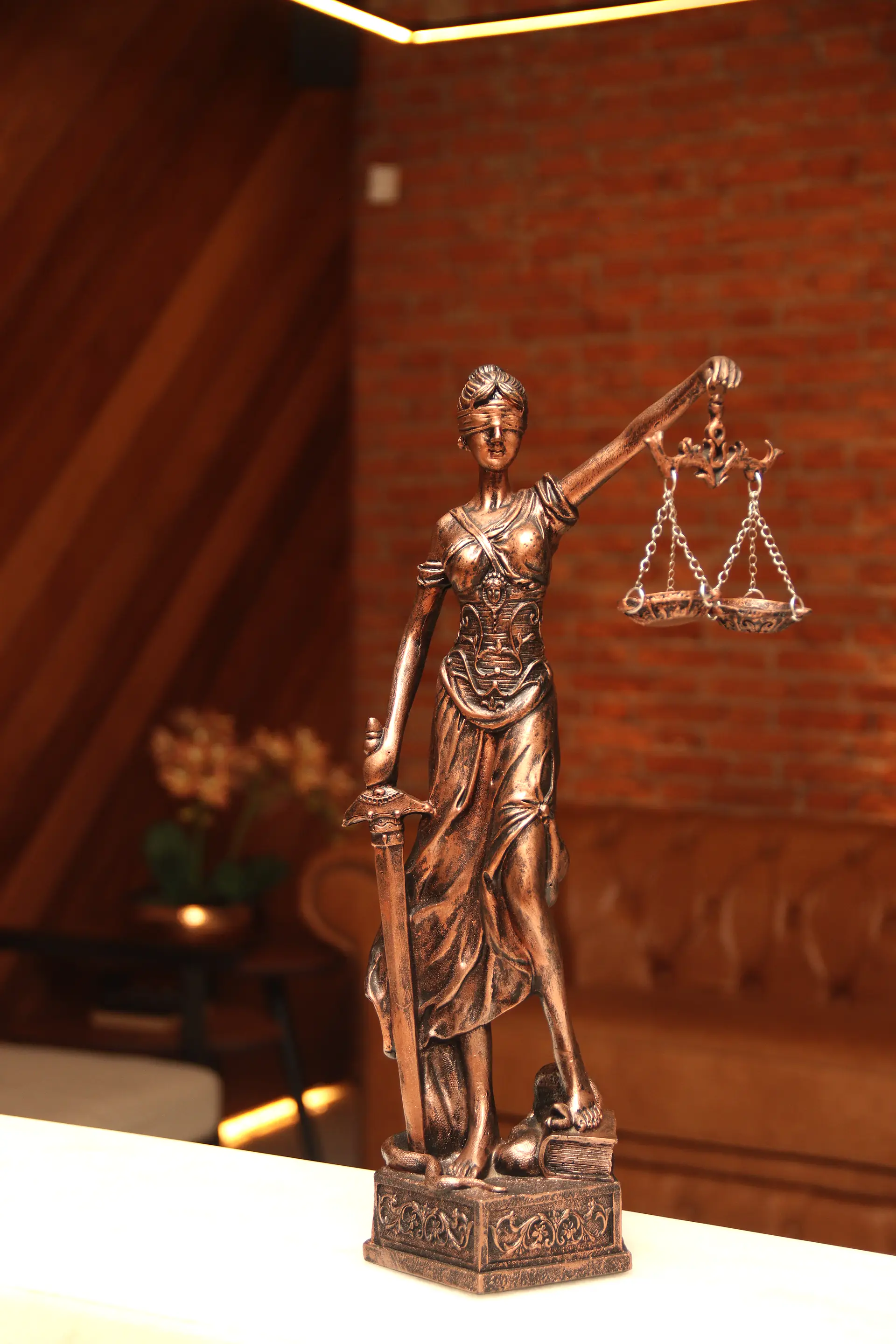 Estatueta de Têmis em bronze segurando a balança da Justiça, em primeiro plano diante da parede de tijolos