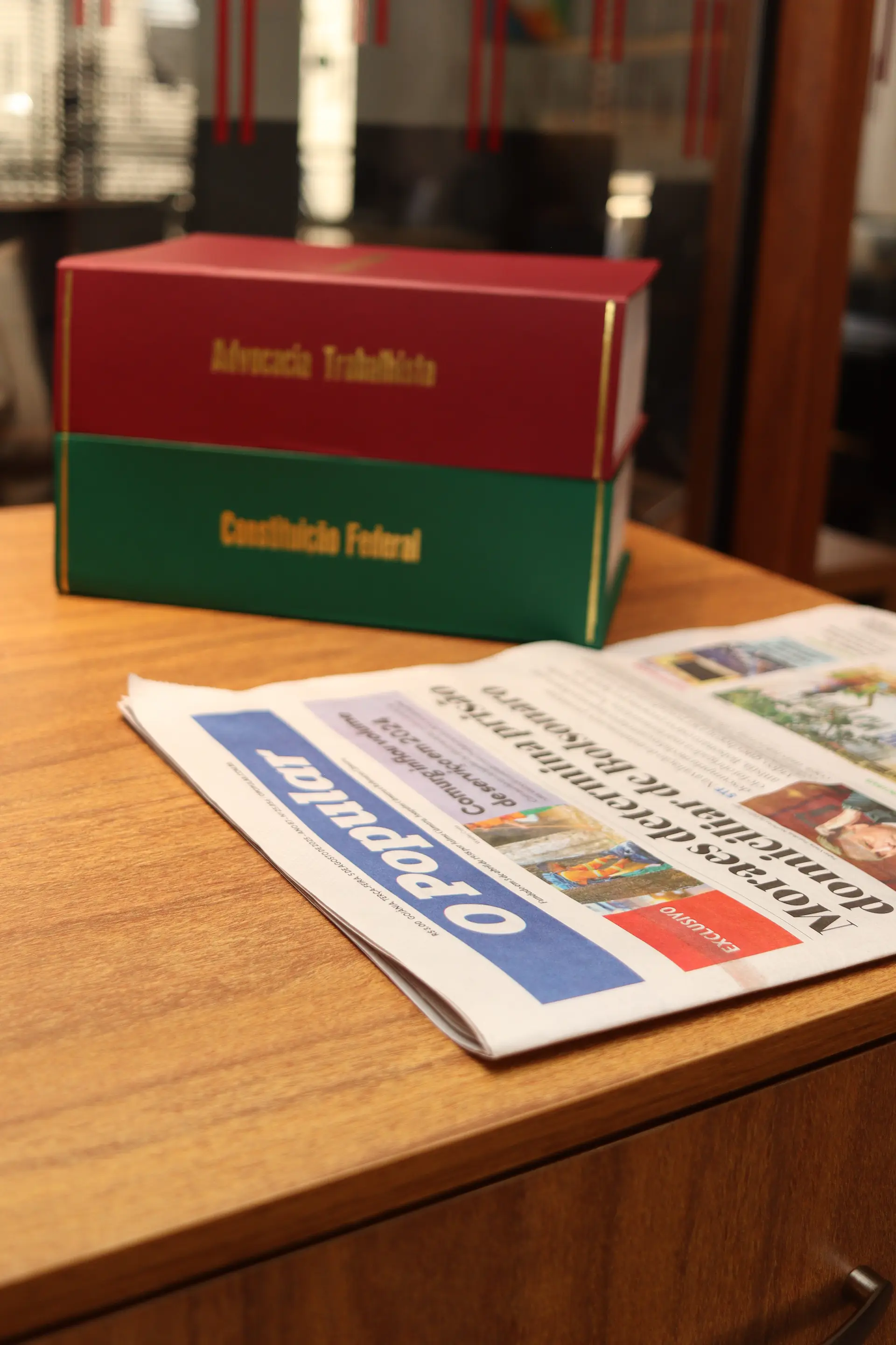 Livros "Advocacia Trabalhista" e "Constituição Federal" empilhados sobre mesa de madeira ao lado de jornal dobrado