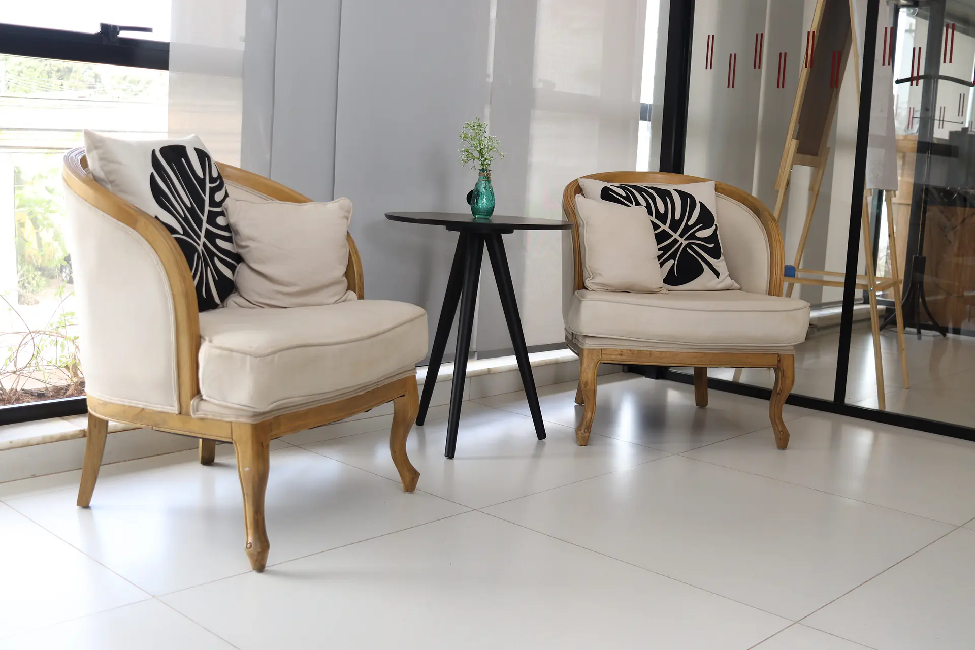 Lounge claro com duas poltronas de linho e madeira entalhada, almofadas com estampa de folhas e mesa lateral preta com vaso verde