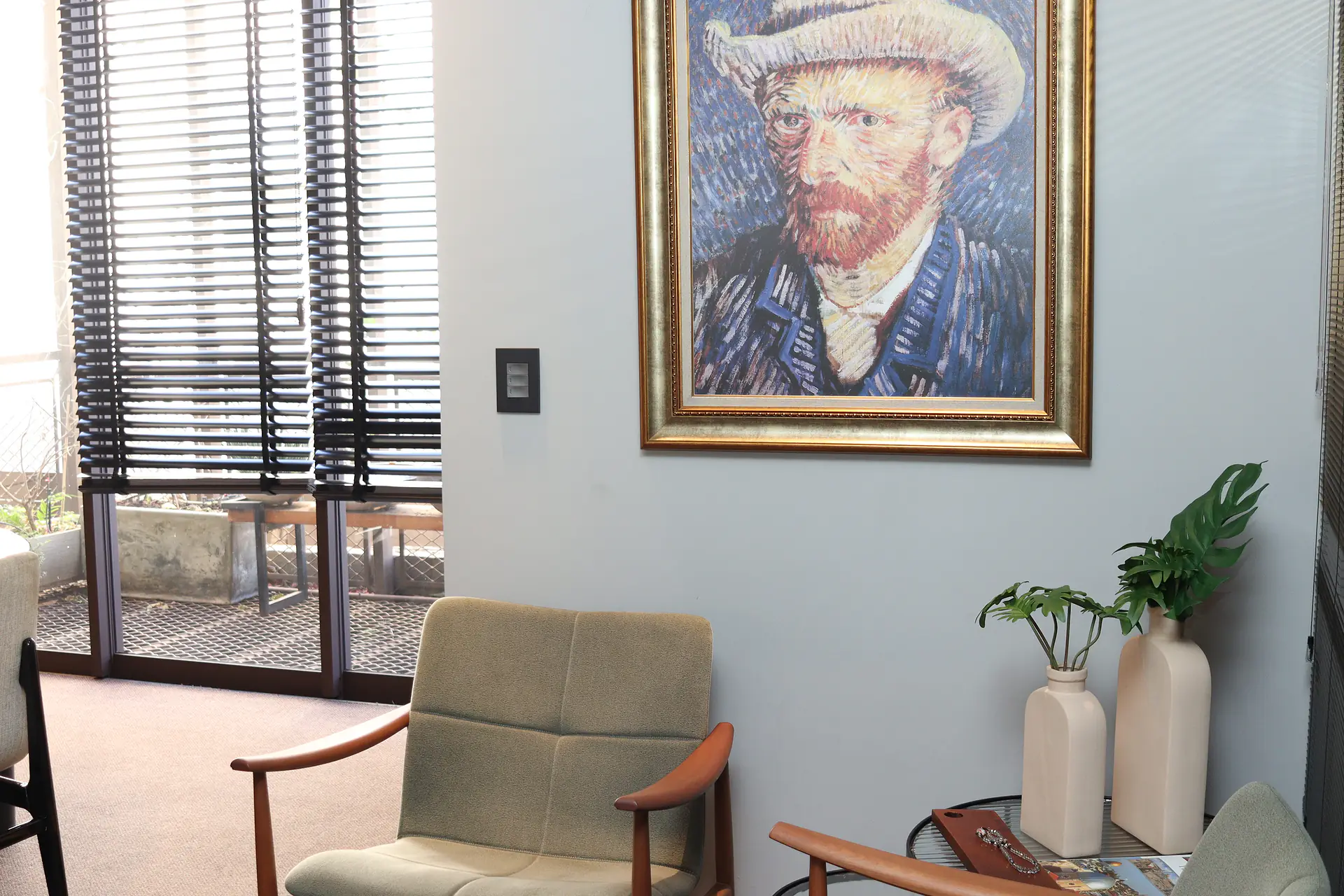 Sala de atendimento com poltronas estofadas em verde-claro, vasos com plantas, autorretrato de Van Gogh em moldura dourada e porta de vidro para a varanda
