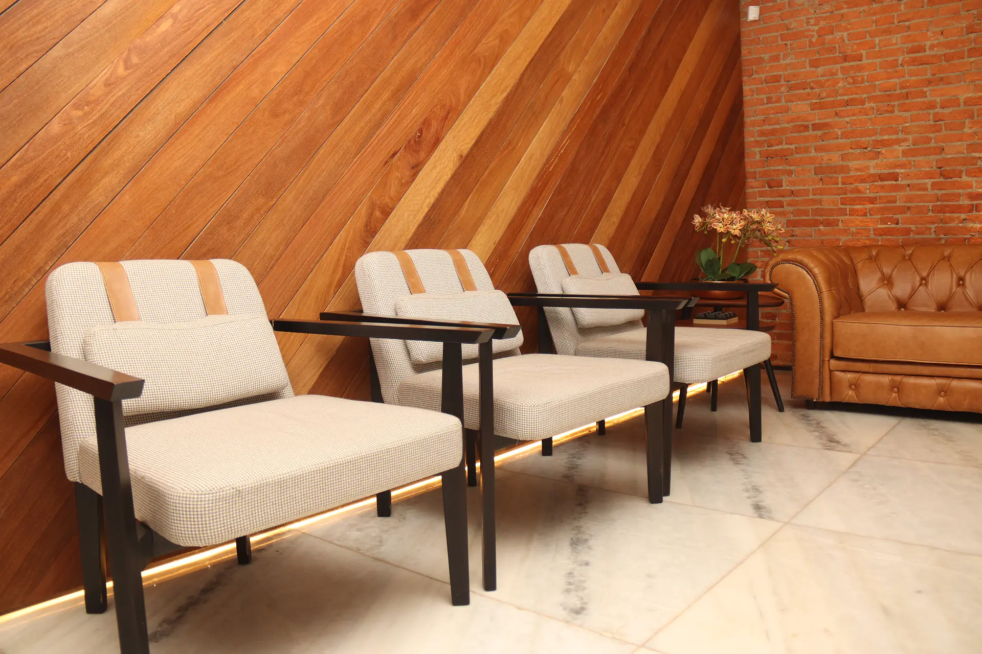 Poltronas estofadas em tecido claro com iluminação indireta no rodapé e sofá de couro ao fundo na sala de espera