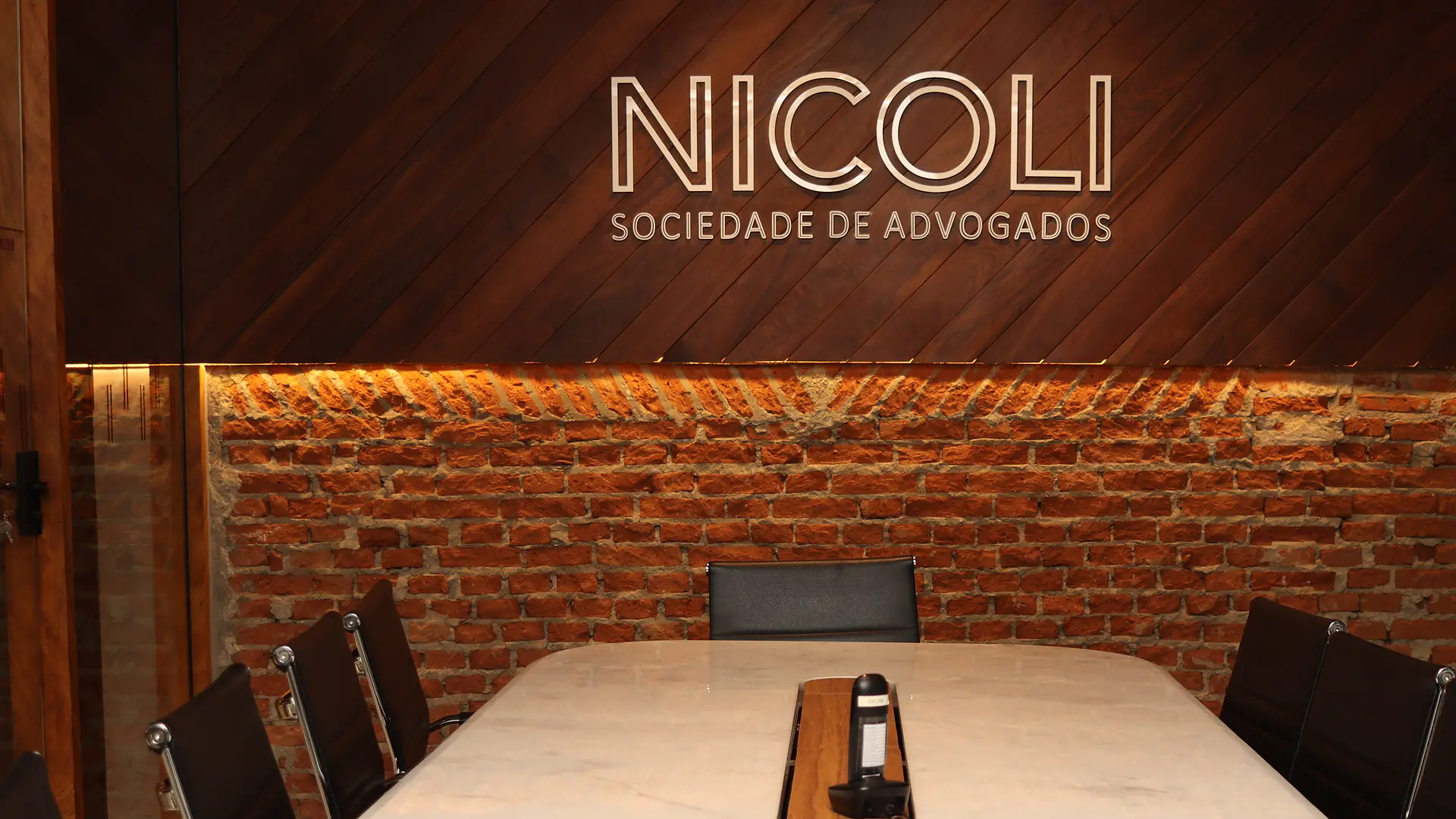 Sala de reuniões com mesa de mármore branco, cadeiras executivas pretas e letreiro "Nicoli Sociedade de Advogados" sobre parede de madeira e tijolos iluminados