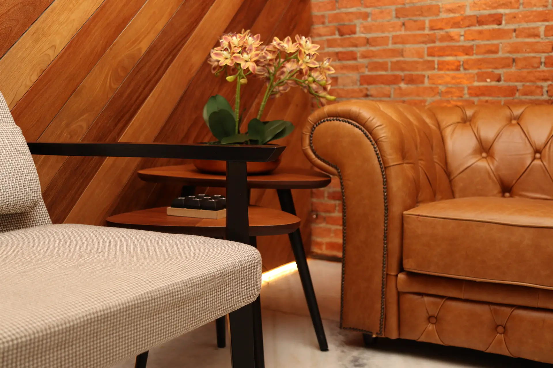 Detalhe do sofá chesterfield em couro caramelo com capitonê, mesa lateral com orquídea e poltrona em tecido pied-de-poule diante da parede de tijolos