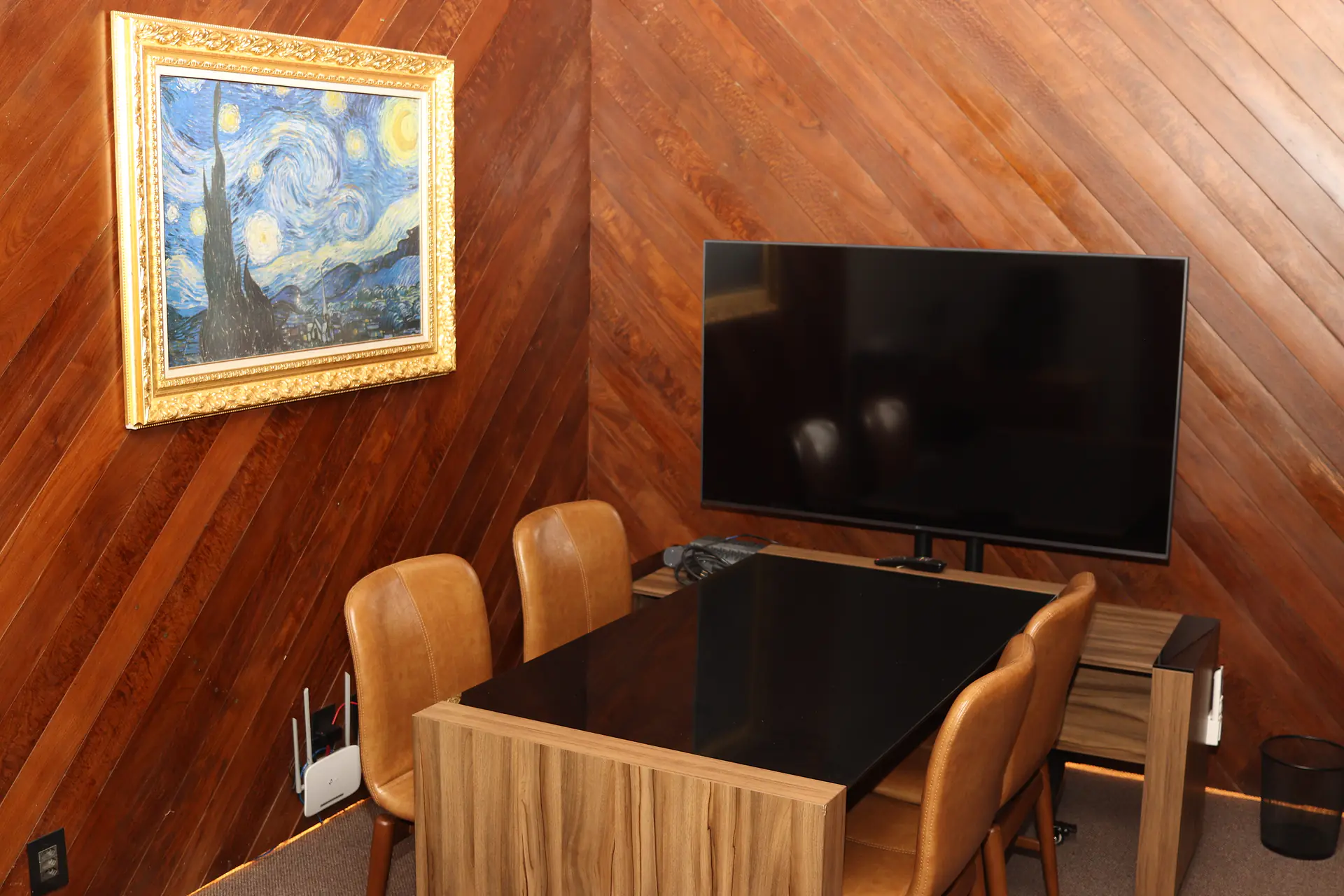 Sala de reunião com mesa de vidro escuro, cadeiras de couro caramelo, televisor e reprodução de "A Noite Estrelada" em parede de madeira
