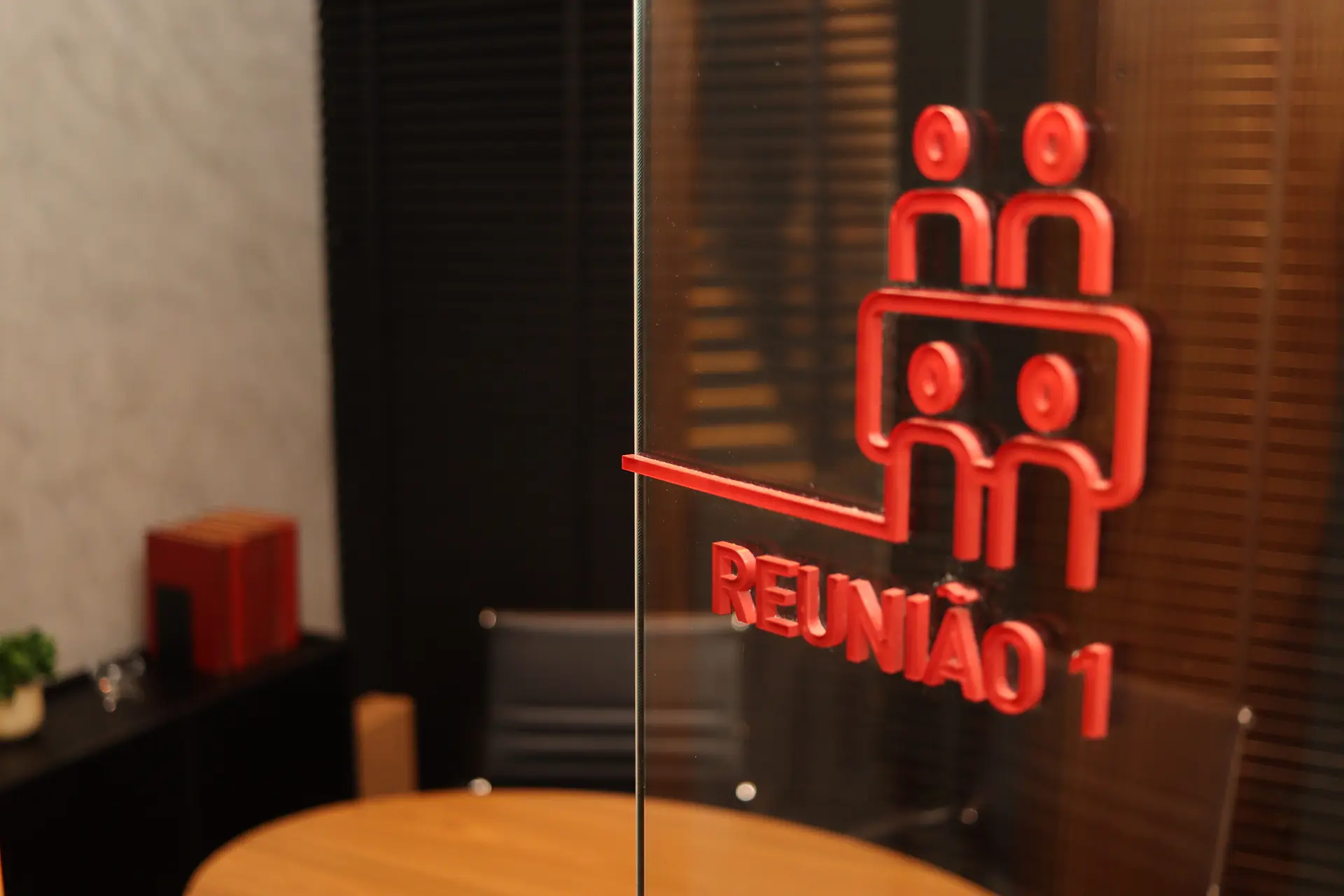 Sinalização em acrílico vermelho "Reunião 1" com ícone de pessoas na porta de vidro, com mesa redonda de madeira desfocada ao fundo